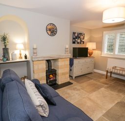 A living room with a blue sofa a fireplace a television on a cabinet and a bench under a window at Dazie Cottage in Broadway