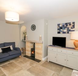 A living room with a blue sofa a small fireplace a wall clock a cabinet with a television and a lamp at Dazie Cottage in Broadway