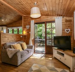 A living room with a couch, cushions, a television on a stand, French doors leading to an outdoor patio with a table and chairs at Scafell Lodge Skiptory Howe 7, White Cross Bay Holiday Park near Windermere