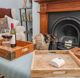 A room with an upholstered chair cushions wooden trays wine bottle and glasses by a fireplace at Anchor Cottage in Whitby