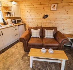 A small living space with a brown leather sofa with two plaid cushions a wooden coffee table with two cups a kitchenette with white cabinets and a window at Robins Nest in Napton-On-The-Hill