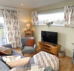 A living room with a gray sofa, an armchair, a wooden TV stand with a TV, a side table with a lamp, patterned curtains, and a small fireplace at The Swift in Wem