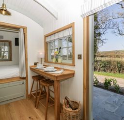 A small dining area with two wooden stools and a built-in bed next to an open door showing an outdoor view at Acorn in Bleadon