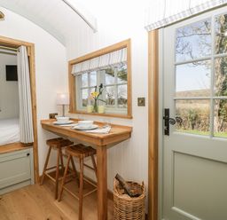 A small dining area with two wooden stools a wooden table a basket with firewood beside a door and a window with flowers at Acorn in Bleadon