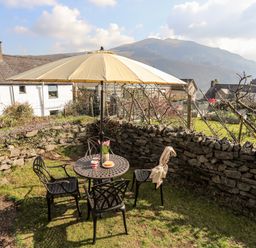 A garden seating area with a round metal table under a large umbrella and four chairs surrounded by a stone wall at 9 Ty Du Road in Llanberis