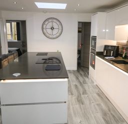 A modern kitchen with a black countertop island and stools a large wall clock and white cabinetry at Sunnydene in Paignton