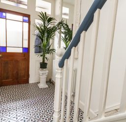 A hallway with patterned tile floor a wooden door with blue stained glass and a potted plant on a white pedestal at ODYSEA in Weymouth