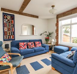 A living room with blue sofas and armchair with patterned cushions a wall hanging and a large window showing plants outside at ODYSEA in Weymouth