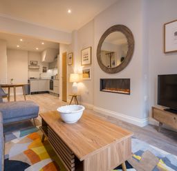 A living room with a gray sofa wooden coffee table a wall-mounted electric fireplace and an open kitchen area at Howe Keld 1 Ground floor in Keswick