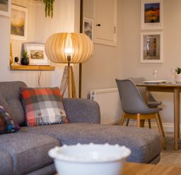 A living room with a gray sofa with patterned cushions a wooden floor lamp and a dining table with chairs at Howe Keld 1 Ground floor in Keswick