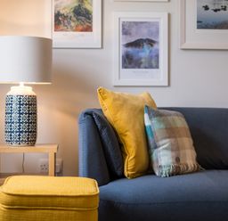 A living room with a blue sofa with two cushions a yellow ottoman a wooden side table with a patterned lamp and three framed pictures on the wall at Howe Keld 6 Second Floor in Keswick