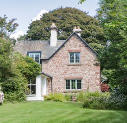 A stone house with green windows surrounded by trees and a lawn at Caldhu Cottage in Holmrook