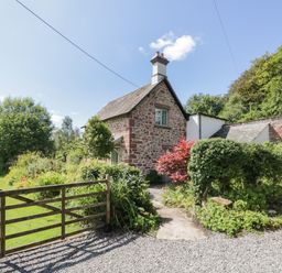 A stone cottage with a chimney surrounded by bushes and trees behind a wooden gate at Caldhu Cottage in Holmrook