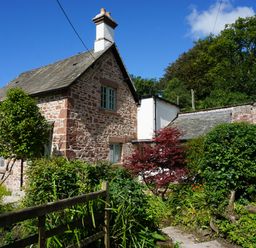A stone cottage with a slate roof surrounded by green bushes and trees at Caldhu Cottage in Holmrook