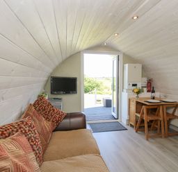 A small living area with a sofa and cushions a wall-mounted TV a wooden dining table with two plates and chairs and an open door leading outside at Cherry Pod in Hookagate near Meole Brace