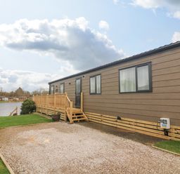 A brown mobile home with windows and a wooden deck near a lake with grass and gravel at Elim South Lakeland Leisure Village near Tewitfield