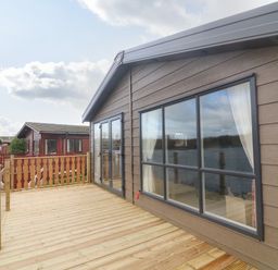 A wooden deck with glass railing attached to a cabin overlooking a body of water at Elim South Lakeland Leisure Village near Tewitfield