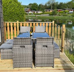 An outdoor deck with a dining table set with chairs and a bottle with glasses overlooking a pond at Elim South Lakeland Leisure Village near Tewitfield