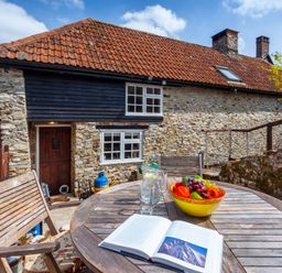 A round wooden table with a fruit bowl open book and glasses on a patio outside a stone cottage at Southcott Farm Cottage in Sheldon
