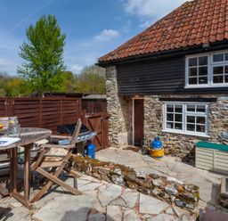 An outdoor patio with wooden table and chairs a barbecue grill and a stone wall next to a stone and wood house at Southcott Farm Cottage in Sheldon