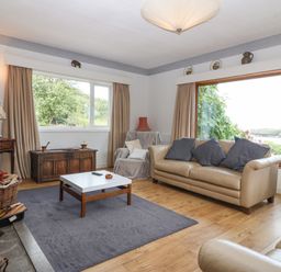 A living room with beige sofas a wooden chest a coffee table and large windows showing greenery and water at The Headland in Llaneilian near Amlwch
