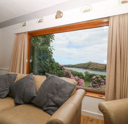 A living room with a beige sofa and gray cushions facing a large window overlooking a garden and water at The Headland in Llaneilian near Amlwch