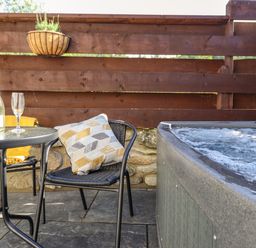 An outdoor patio with a bubbling hot tub a black metal table with two glasses and a bottle and two black chairs one with a patterned cushion at Glyndwr in Betws-Y-Coed