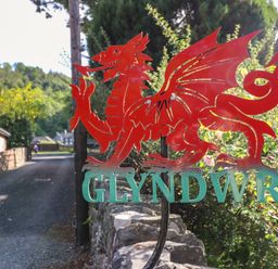 A red dragon metal sign with the word glyndwr on a stone wall by a gravel road with houses and trees in the background at Glyndwr in Betws-Y-Coed