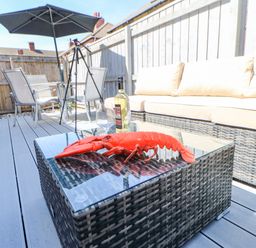 An outdoor patio with a wicker sofa a glass table with a lobster a bottle of wine and a glass a dining table with chairs and an umbrella at Lobster Pot Retreat in Newbiggin-By-The-Sea