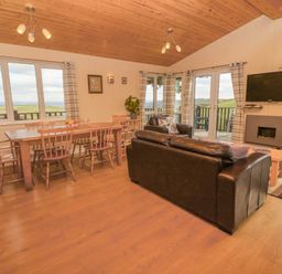 A living room with a wooden dining table and chairs a black leather sofa a mounted TV and windows showing a countryside view at Lodge 23 in Torpoint