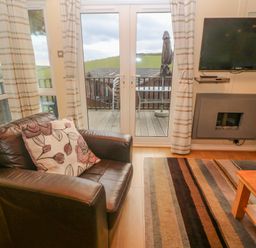 A living room with a leather armchair and floral cushion next to glass doors with striped curtains a wall-mounted TV and fireplace and a striped rug on the floor at Lodge 23 in Torpoint