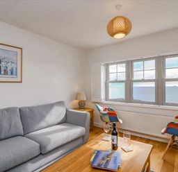 A living room with a grey sofa two colorful chairs a wooden table with wine glasses and books and a window with a sea view at Beach Retreat in Weymouth