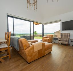 A living room with a brown sofa set, wooden dining table with chairs, large windows, and a wall mounted TV at Owl Lodge in Wedmore