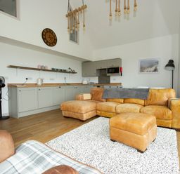 A living room with a tan leather sofa a matching ottoman a white patterned rug and a kitchen with gray cabinets at Starling Lodge in Wedmore