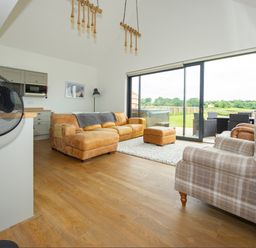 A living room with brown leather sofa and plaid armchair near large sliding glass doors showing outdoor patio at Starling Lodge in Wedmore
