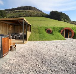 An outdoor area with a hot tub and covered seating next to a grass-covered hobbit-style house with circular windows and door at Nant yr Esgair Mallwyd near Dinas Mawddwy