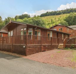 A wooden lodge with a fenced porch surrounded by trees and hills at Kirkstone Lodge in Limefitt Park near Windermere
