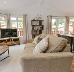 A living room with a beige sofa, wooden coffee table, shelves, TV on a wooden stand, dining table with chairs and wine glasses at Kirkstone Lodge in Limefitt Park near Windermere