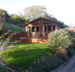 A wooden cabin with a fenced porch surrounded by grass and bushes at 23 Bossiney Bay in Tintagel