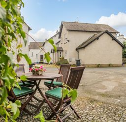 An outdoor seating area with a round wooden table and four chairs with green cushions on a cobblestone surface with white houses in the background at Columbine Cottage in Hawkshead