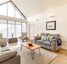 A living room with two grey sofas two beige chairs a wooden coffee table and large windows at Forget Me Not Lodge Bamburgh