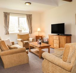 A living room with beige sofas a wooden coffee table a wooden TV cabinet a window with curtains and a small lamp at Tilly Mint Cottage Nr Ullswater