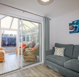 A living room with a gray sofa and patterned pillows next to a conservatory with wicker chairs and a garden outside at Chesil Retreat in Weymouth