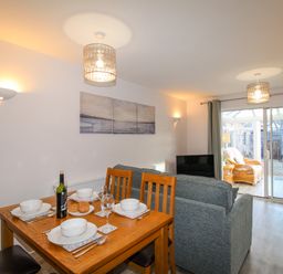 A dining area with a wooden table set for four next to a living room with a grey sofa and sliding glass doors leading to a room with wicker chairs at Chesil Retreat in Weymouth