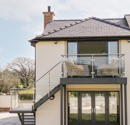 A house with a balcony and outdoor chairs with trees in the background at The Annexe in Llanfairpwllgwyngyll