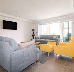A living room with grey sofas yellow armchairs a wall-mounted TV and large windows at 30 Cliff Edge in Newquay