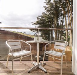 A balcony with two metal chairs and a round table with two mugs overlooking trees and a wooden fence at 11 Towan Valley in Porthtowan