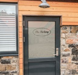 A black door with frosted glass and the words The Bakery on it next to a window with white shutters on a stone and wood exterior at The Bakery in Middleton near Belford