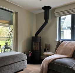 A living room with a wood stove next to a window and a gray armchair with a blanket at Primrose Hellandbridge near Bodmin