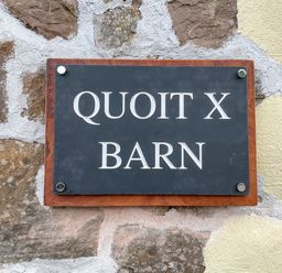 A stone wall with a black and brown sign reading Quoit X Barn